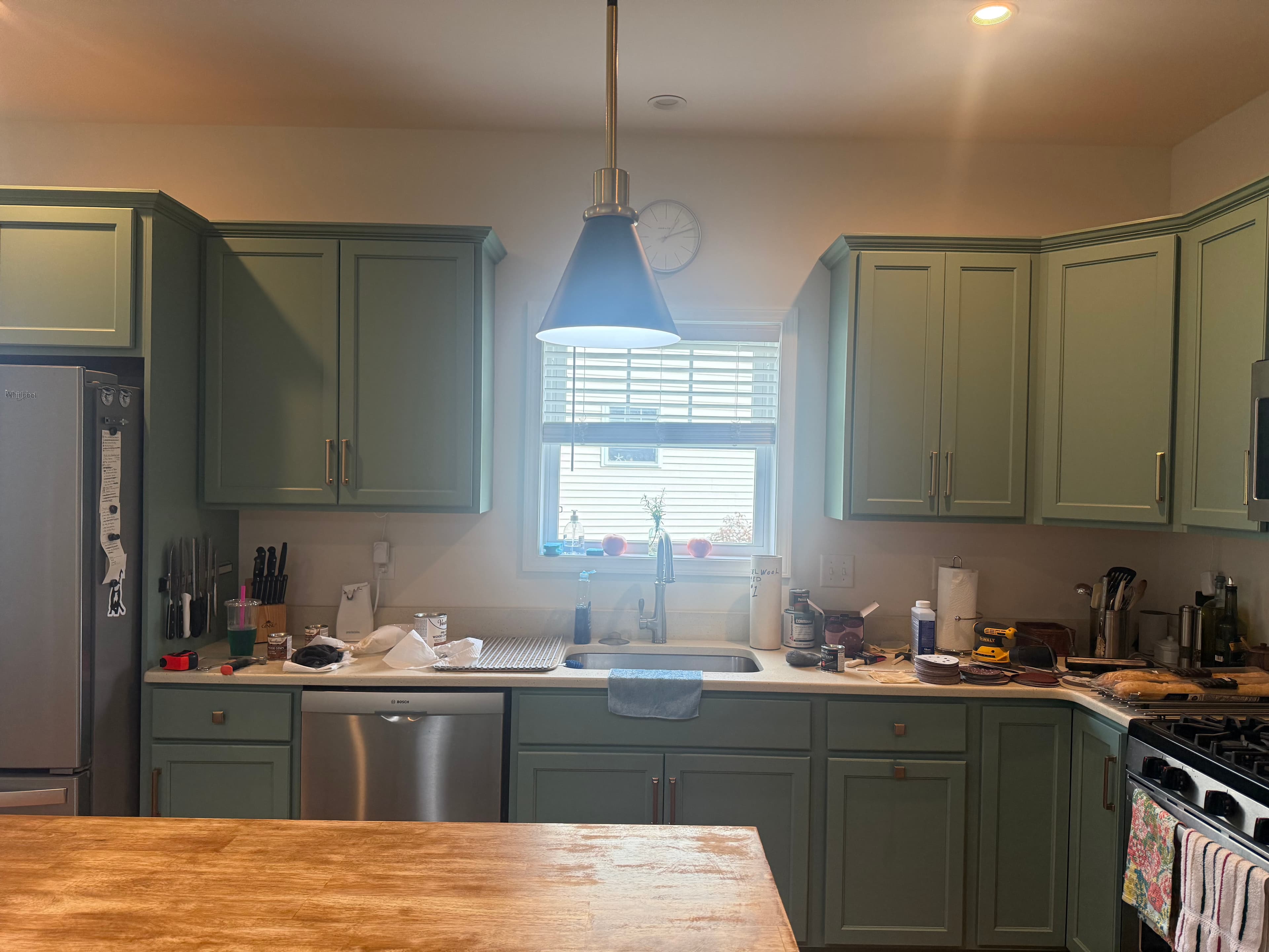 Sage green kitchen cabinets and stainless steel appliances with a cluttered countertop under a window.