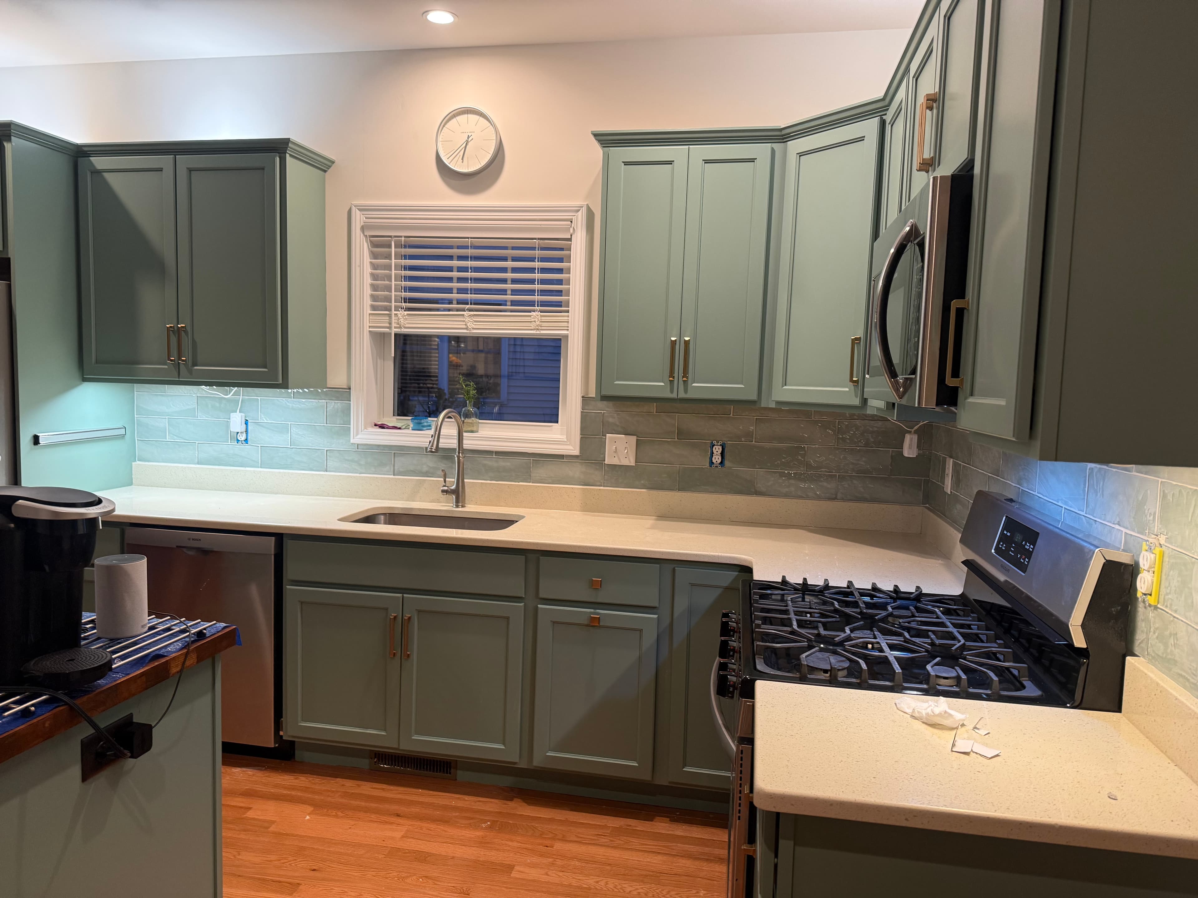 Modern kitchen with sage green cabinets, gold hardware, white countertops, and blue tile backsplash.